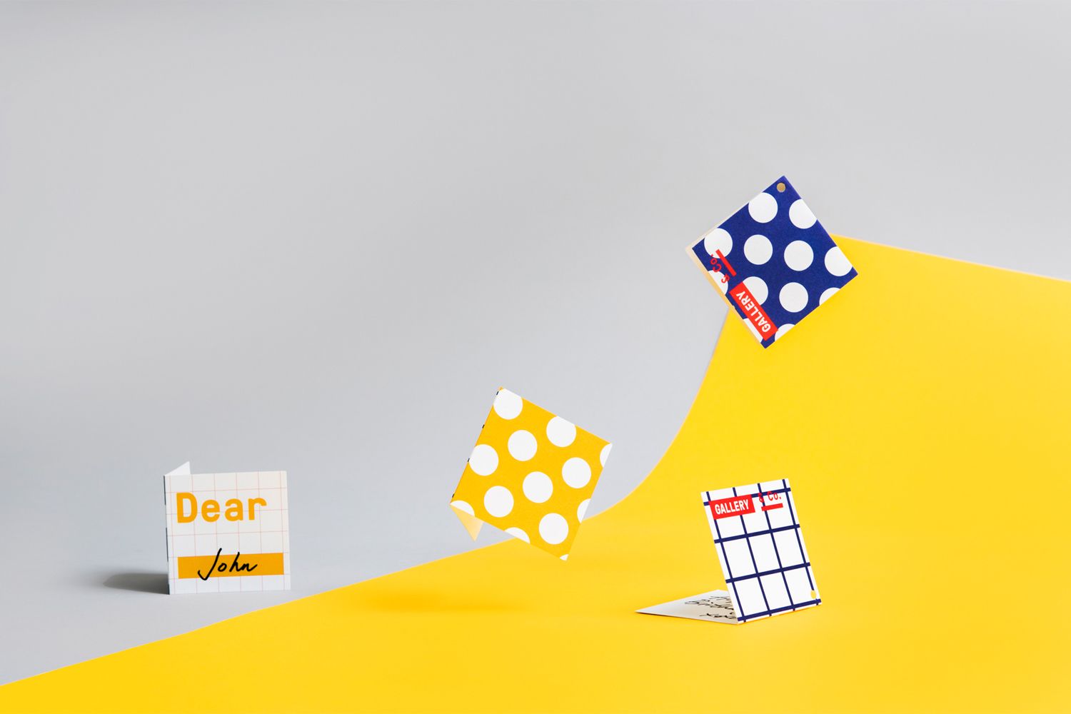 Four folded paper cards with colorful polka dot and grid patterns floating above a bright yellow and grey studio backdrop.