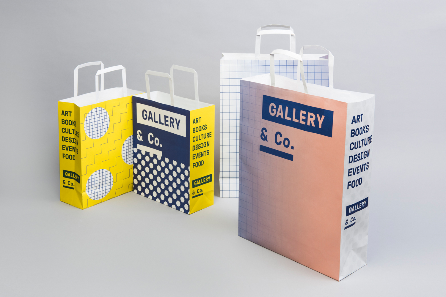 Four branded paper shopping bags with geometric and gradient designs arranged on a grey seamless studio surface.