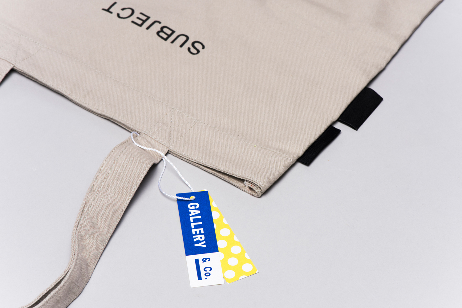 Beige canvas tote bag with black text subject and branded tags resting on a neutral light grey surface