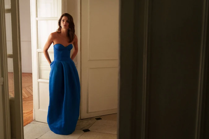 Model Isabeli Fontana wearing a blue strapless gown standing in an apartment interior for BCBGMAXAZRIA Fall 2025 campaign.
