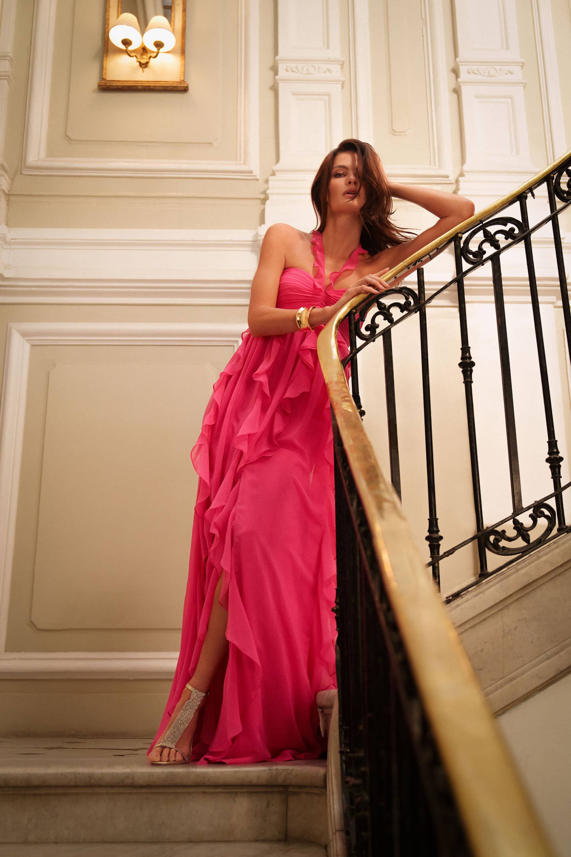 Model Isabeli Fontana in a vibrant pink gown leaning on an ornate staircase railing for BCBGMAXAZRIA Fall 2025 campaign.