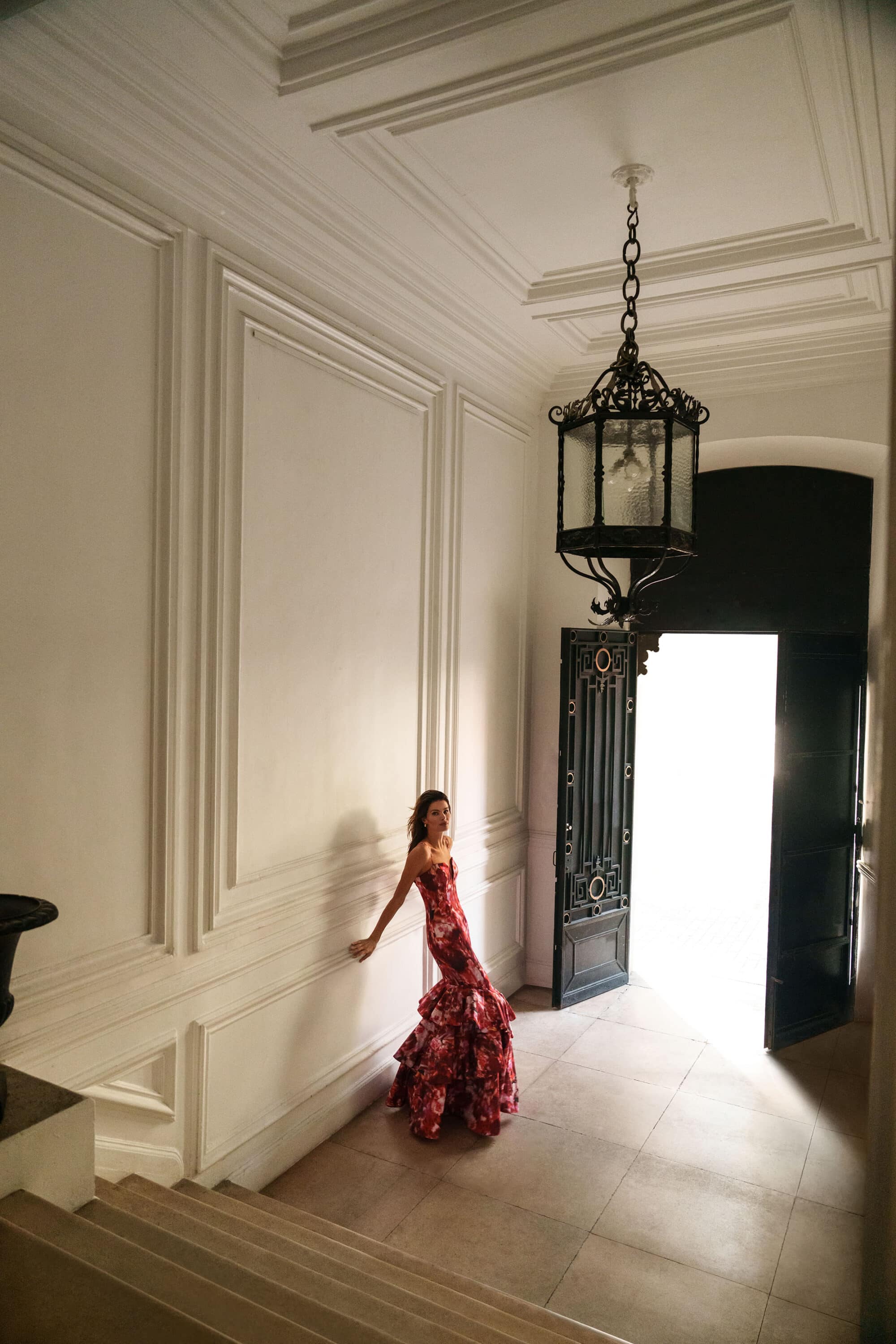 Model Isabeli Fontana in a patterned red gown posing in a grand hall with high ceilings and a pendant light for BCBGMAXAZRIA.