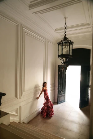Model Isabeli Fontana in a patterned red gown posing in a grand hall with high ceilings and a pendant light for BCBGMAXAZRIA.