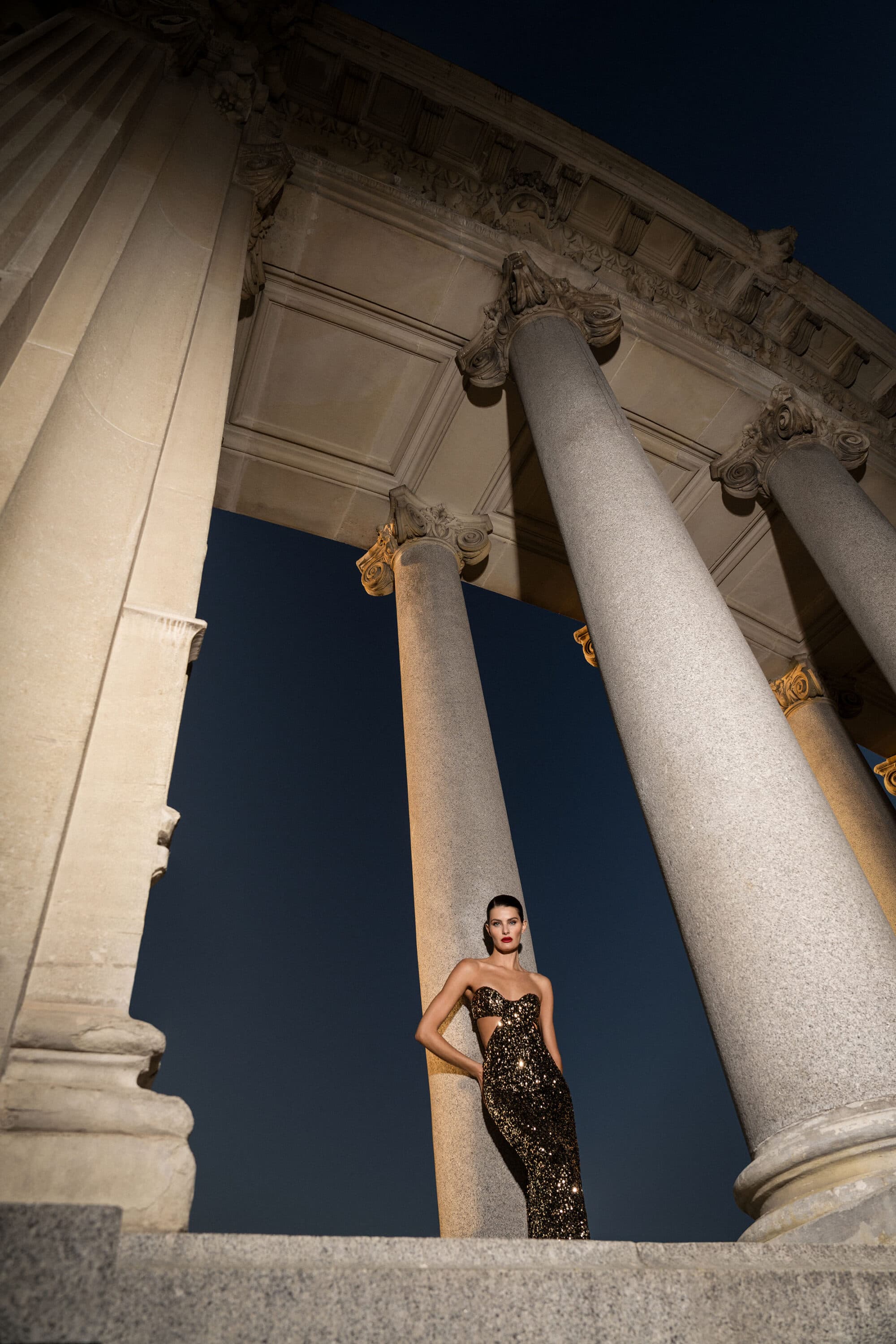 Model wearing a sequined gown leaning against a classical stone column at night for the BCBGMAXAZRIA fall 2025 campaign.