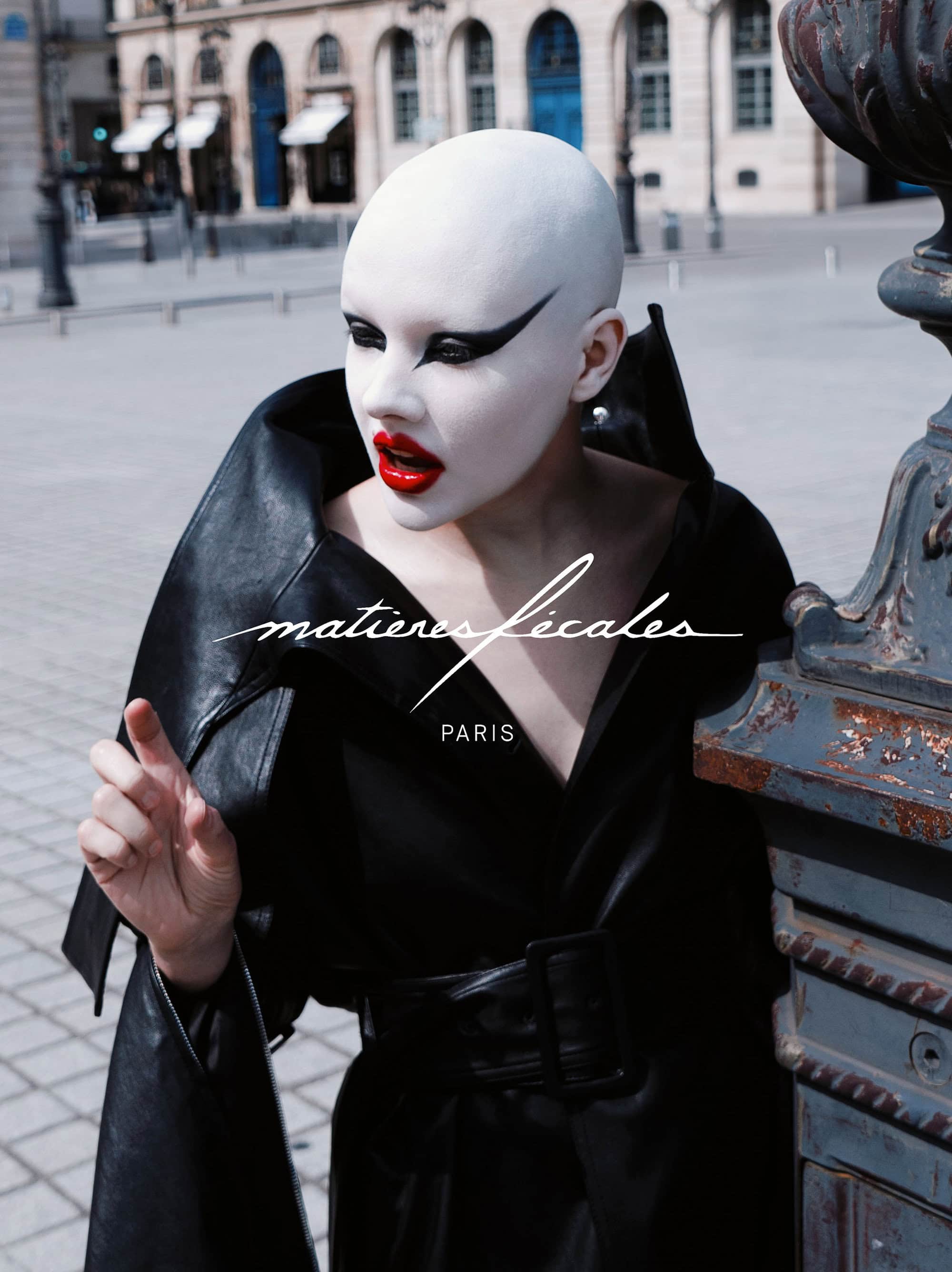 Model with shaved white head and dramatic makeup in black leather jacket posing outdoors on a public square in Paris