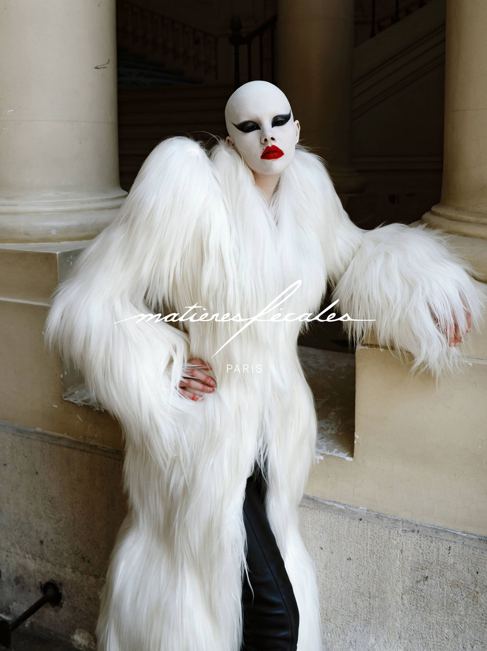 Model wearing dramatic white faux fur coat and bold makeup posing against classical stone pillars outdoors.