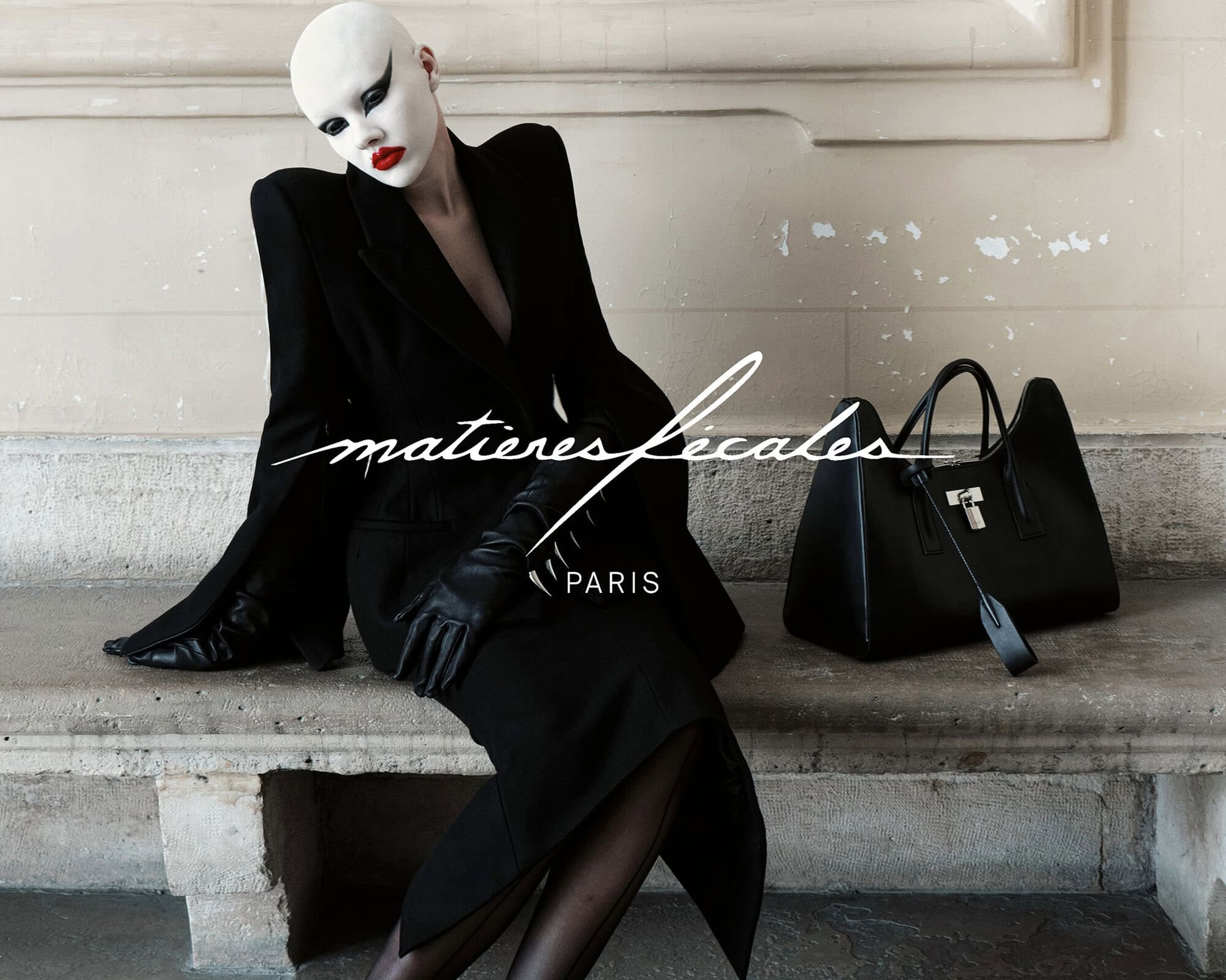 Model in avant-garde makeup and black blazer sitting on a stone ledge next to a black leather handbag