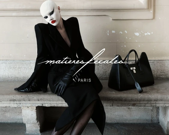 Model in avant-garde makeup and black blazer sitting on a stone ledge next to a black leather handbag