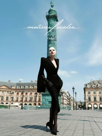 Model wearing a structured black suit and heels standing in a grand public square with a tall column in the background.