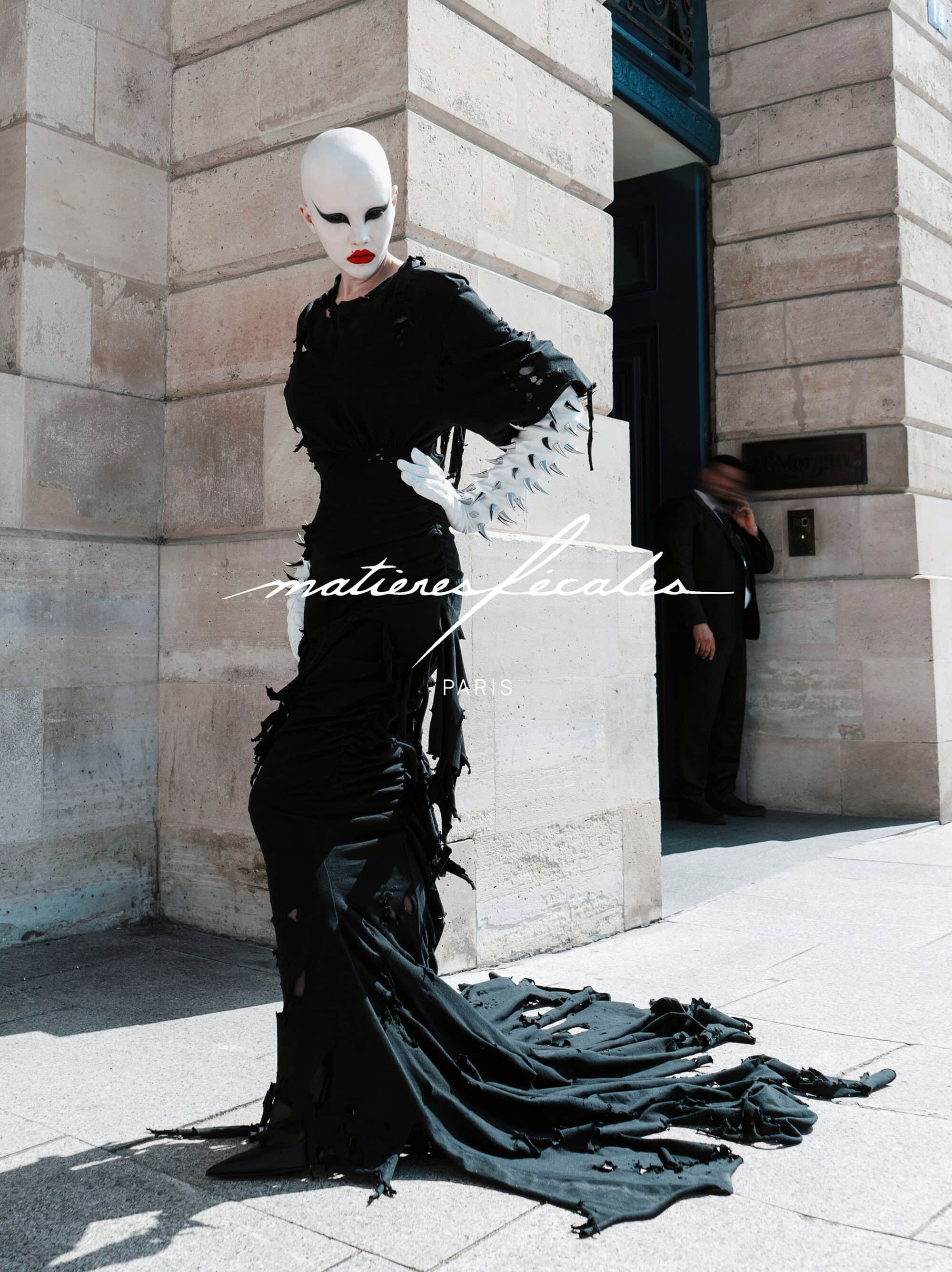 Model in avant-garde black distressed gown with white gloves standing outside classical stone building exterior