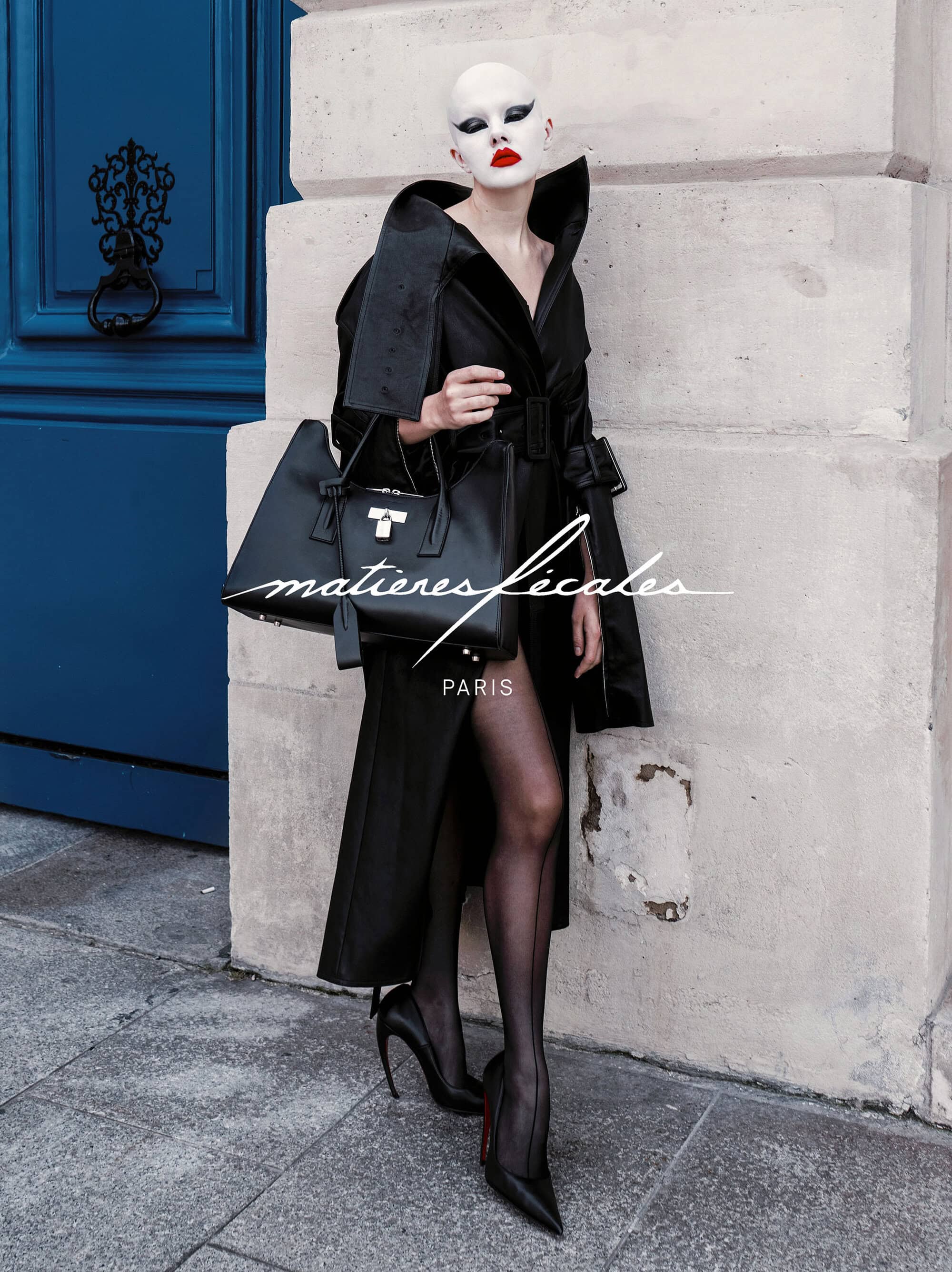 Model in black coat holding a handbag standing against a stone building exterior next to a blue door