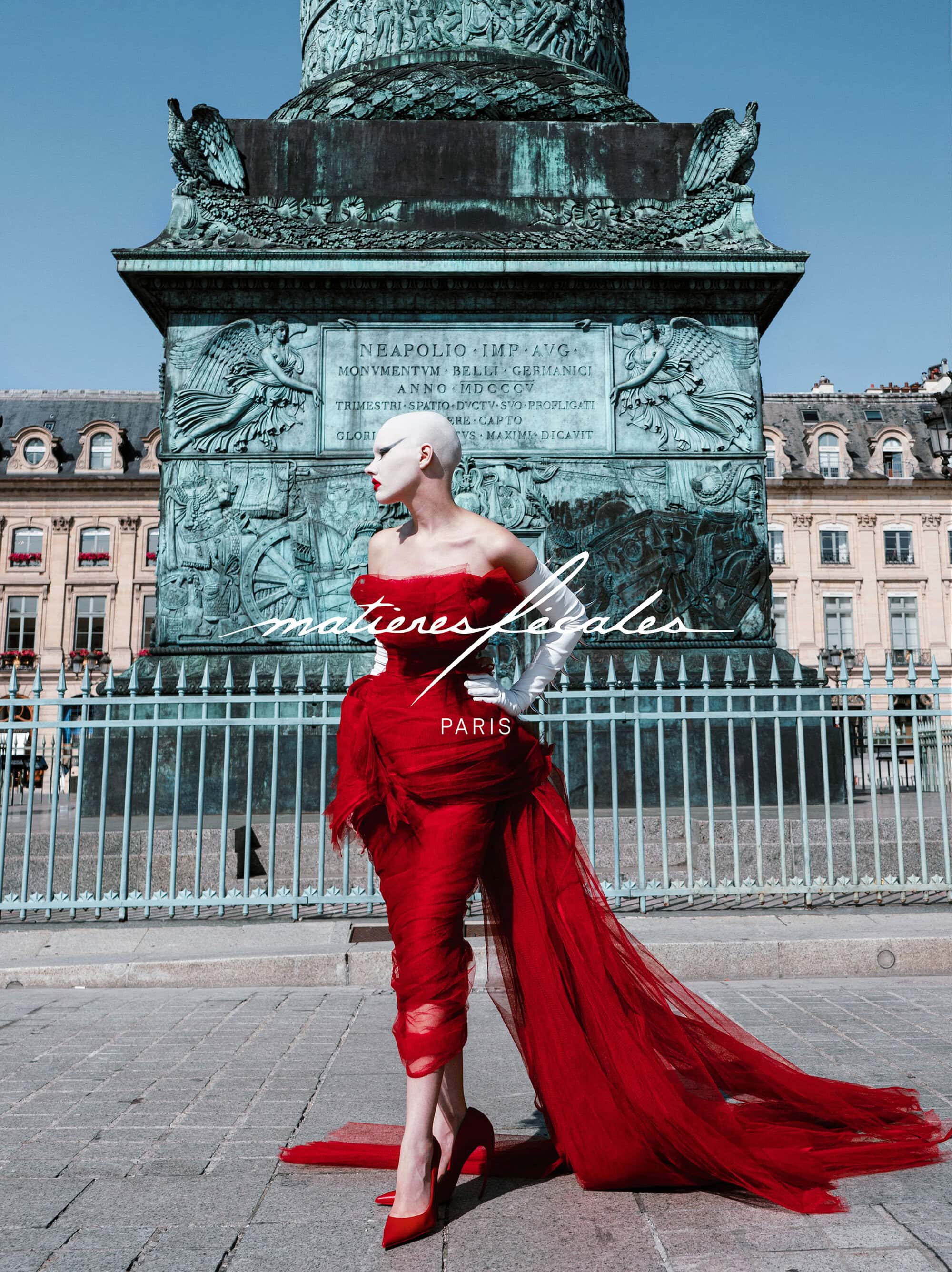 Model with shaved head wearing a dramatic red tulle dress and white gloves standing before the Vendôme column in Paris