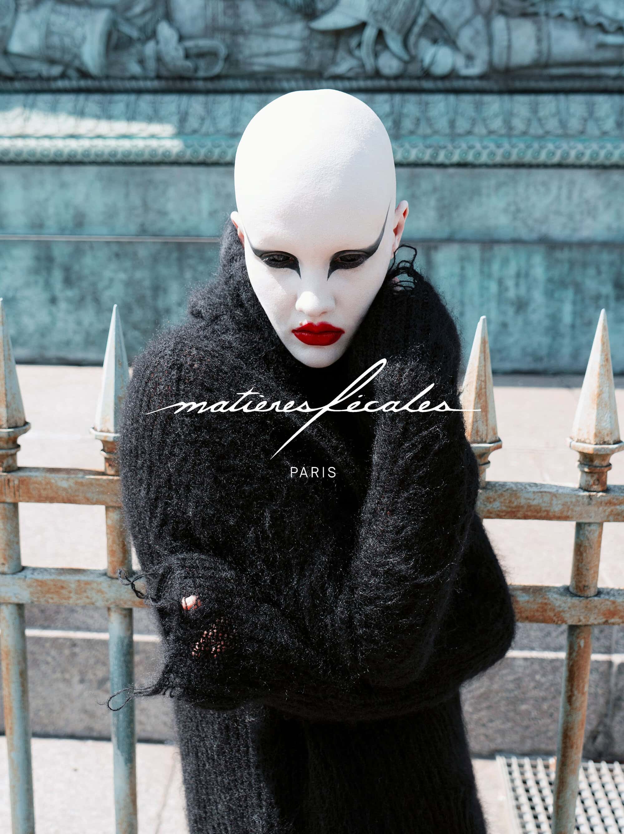 Model with painted white face and black eye makeup wearing a black fuzzy turtleneck sweater behind iron spikes