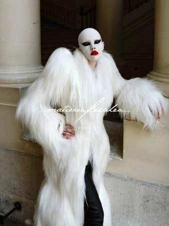 Model wearing dramatic white faux fur coat and bold makeup posing against classical stone pillars outdoors.