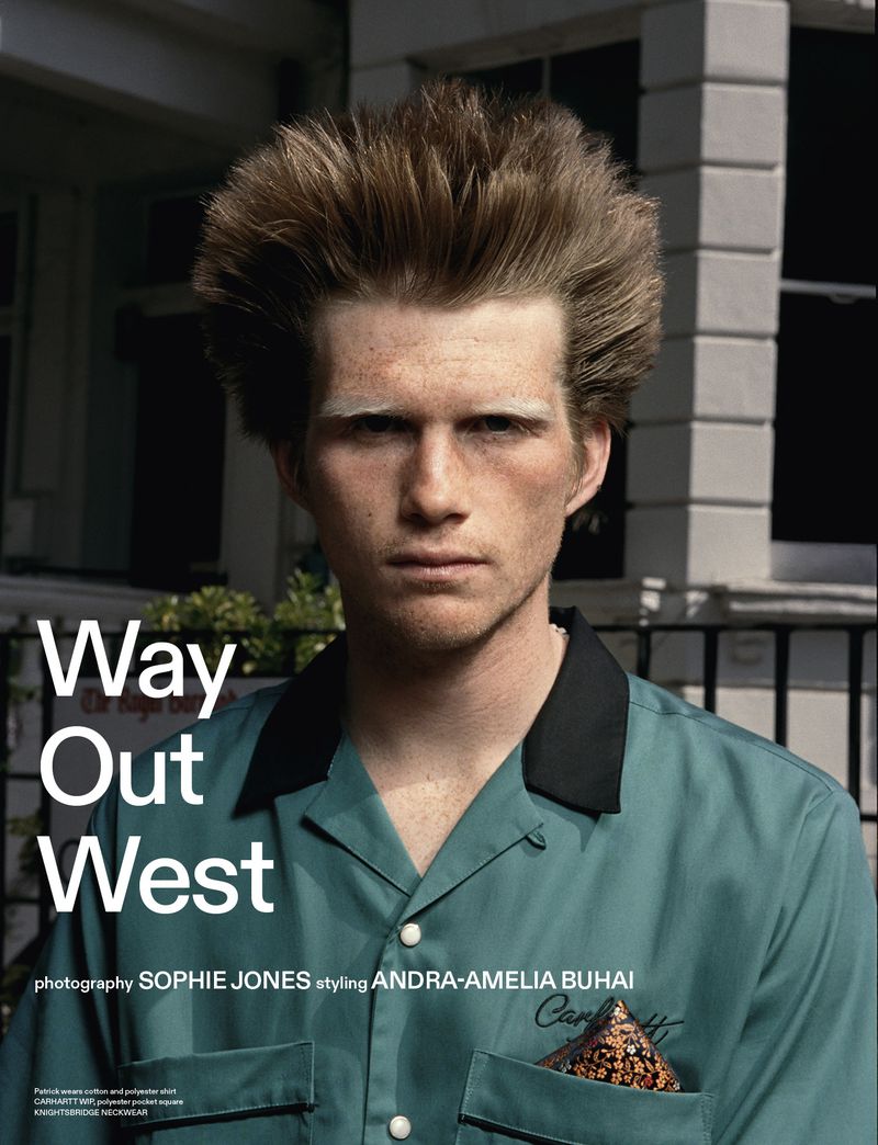 Portrait of a light-skinned adult male with tall hairstyle wearing a teal shirt with black collar standing in front of an urban building.