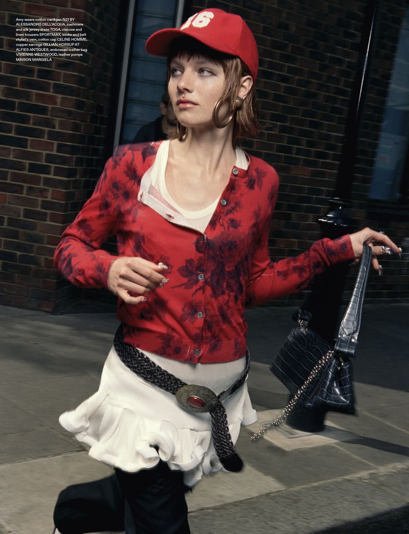 Young adult woman in casual streetwear with red cap poses on a city sidewalk near a brick building with a black purse. Fashion editorial.