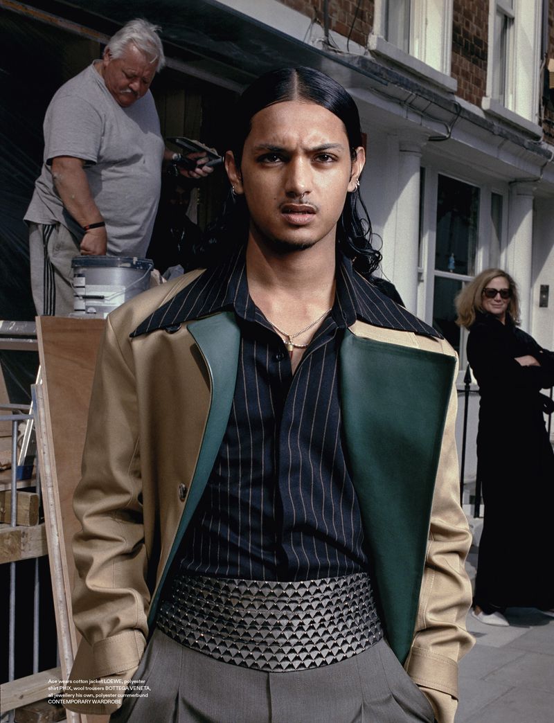 Fashion editorial shot of a young man in a striped shirt, statement belt and jacket, with crew and building background in daylight.