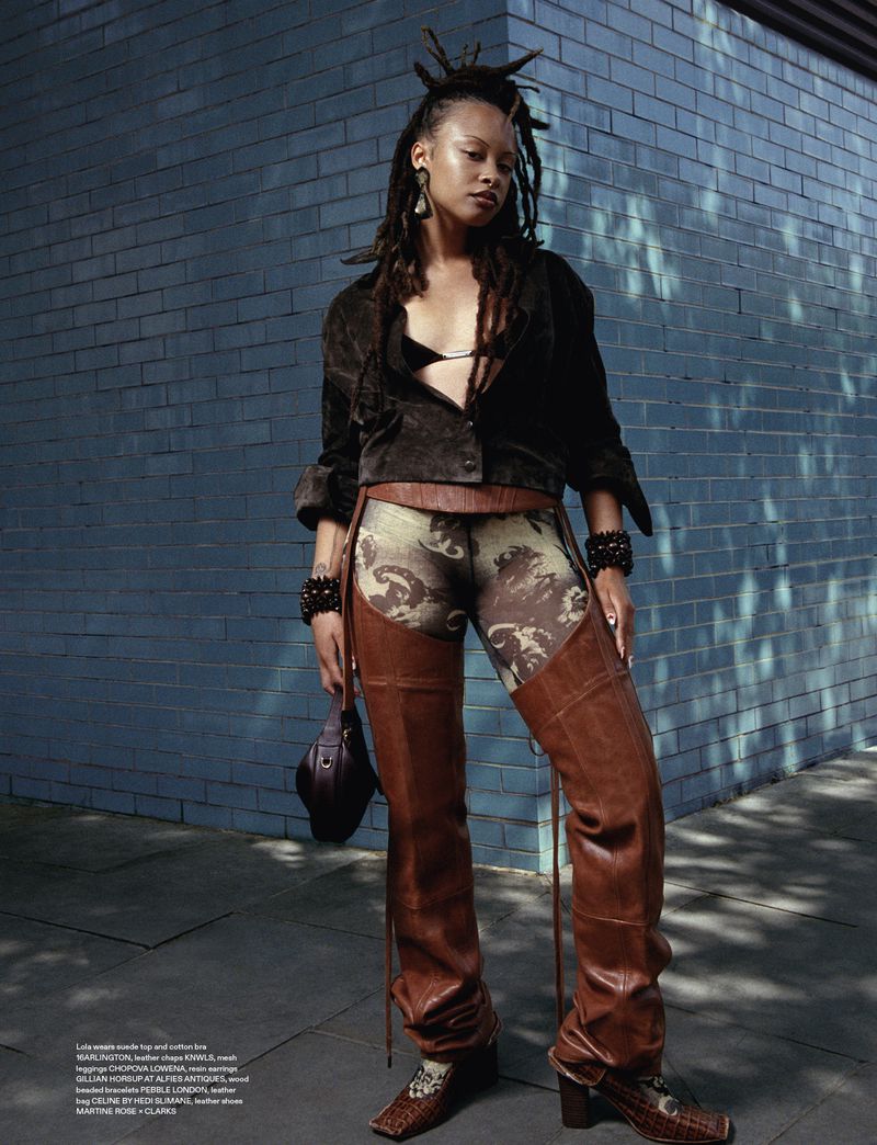 A woman with dreadlocks poses in leather chaps and a suede jacket against a blue brick wall in an outdoor fashion shot.