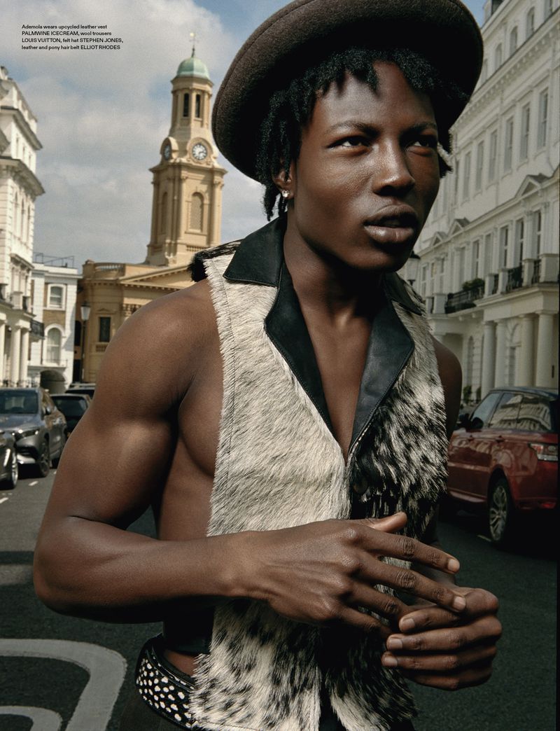 Masculine-presenting person in a sleeveless top stands on a city street near cars and a church clock tower in the background.