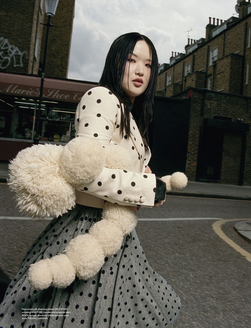 A young woman in a polka dot jacket and skirt strides down a city street holding a stuffed toy, showcasing a blend of fashion and whimsy.