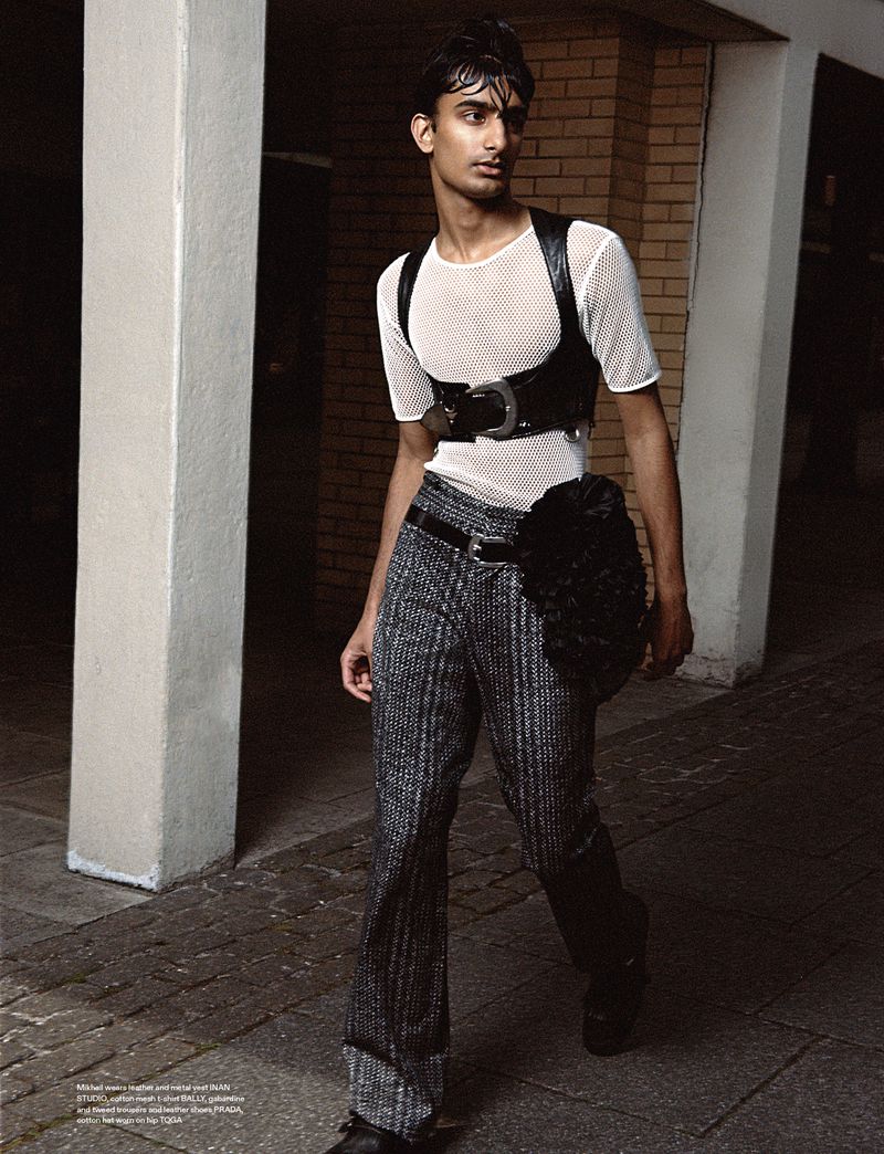 Fashion shot of a young adult male with dark hair, wearing a net shirt and black leather harness, posing on a city sidewalk.