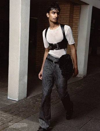 Fashion shot of a young adult male with dark hair, wearing a net shirt and black leather harness, posing on a city sidewalk.
