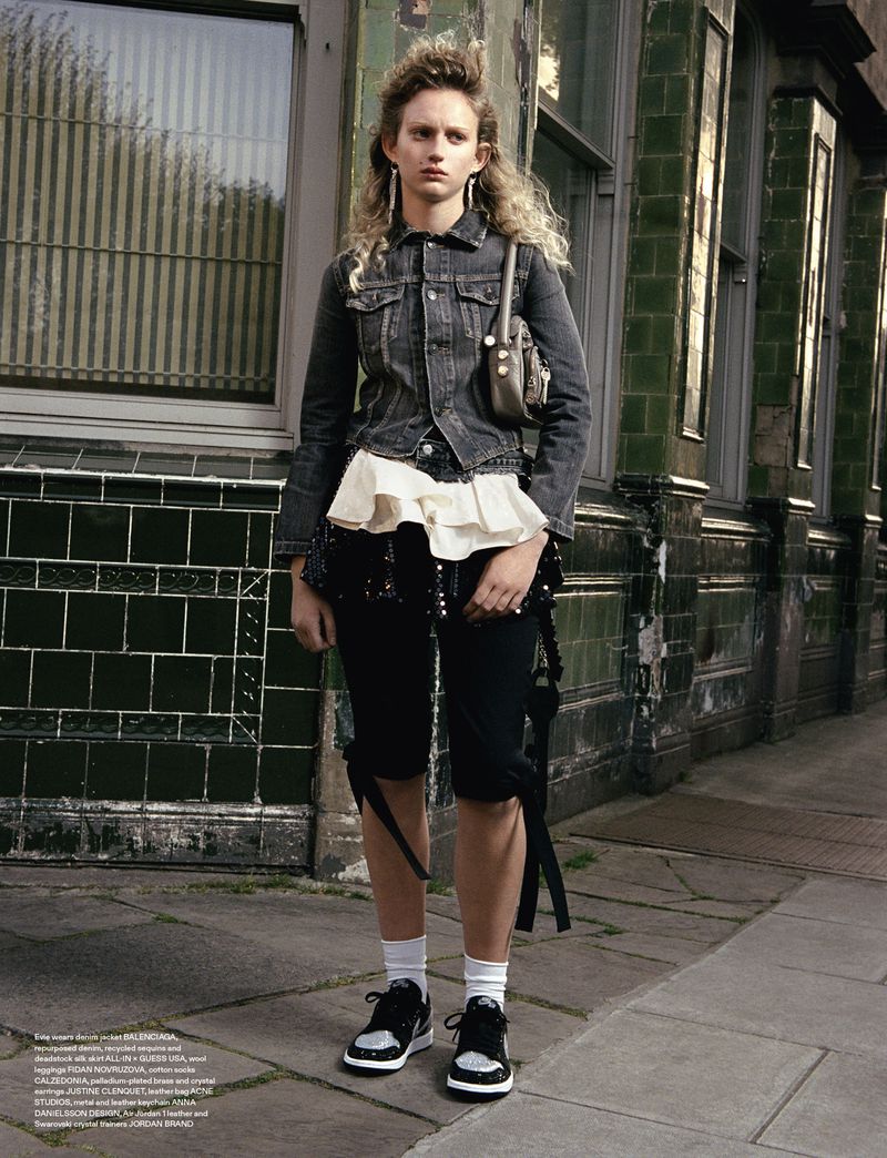Woman in denim jacket and shorts stands on city street near green tile building, accessorized with small purse and black and white sneakers.