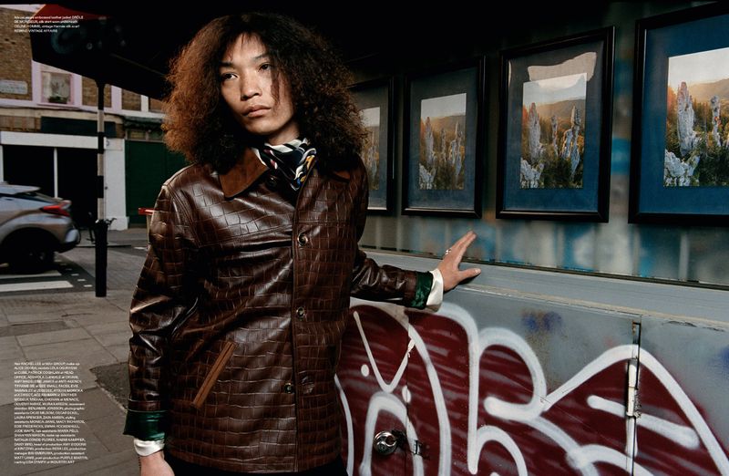 Stylish adult with curly hair in brown leather jacket poses on urban street with graffiti art and framed landscape images in background.