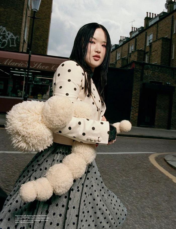Asian woman wearing a polka dot jacket and skirt carrying a fluffy cream plush object on a city street.