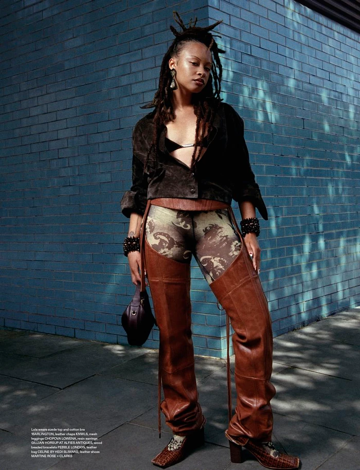 Full-length fashion portrait of a model with dreadlocks wearing brown leather chaps and a dark jacket against a blue brick wall
