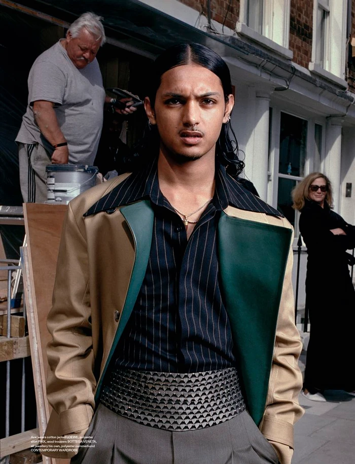 Young man with long dark hair wearing a beige and green color-blocked jacket over a pinstripe shirt and grey trousers on a sunny street.