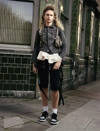 Model with blonde curly hair wearing a dark denim jacket and white rffled blouse standing on a city street next to a tiled building