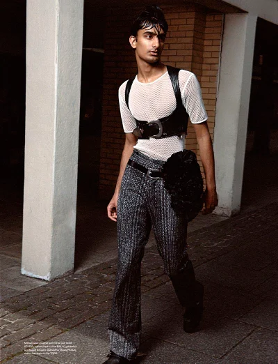 A South Asian male model wearing a white mesh top, black harness, and patterned trousers walking outdoors near concrete pillars