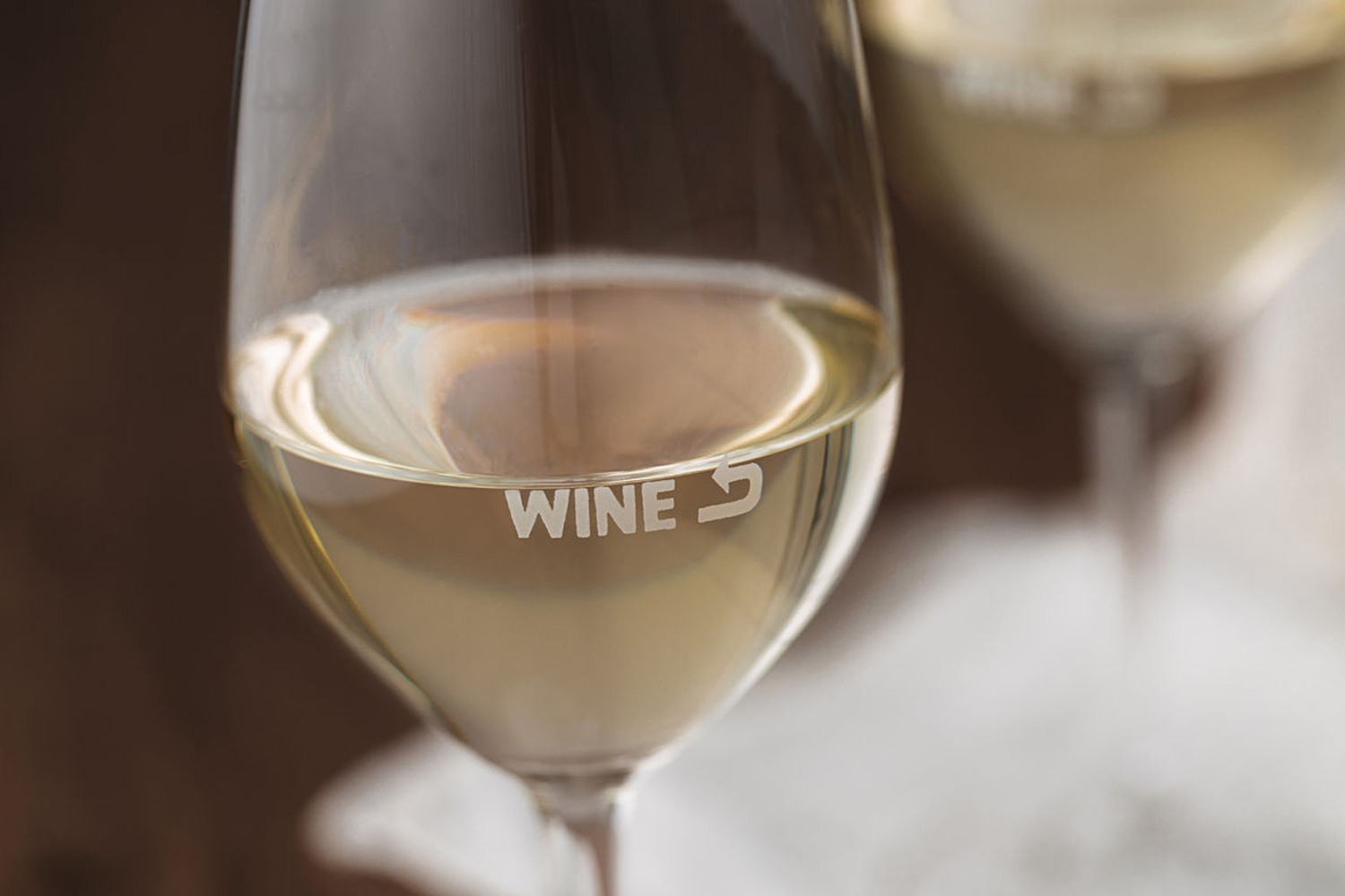 Close-up of a wine glass filled with white wine, featuring branded typography on the glass surface against a neutral background.