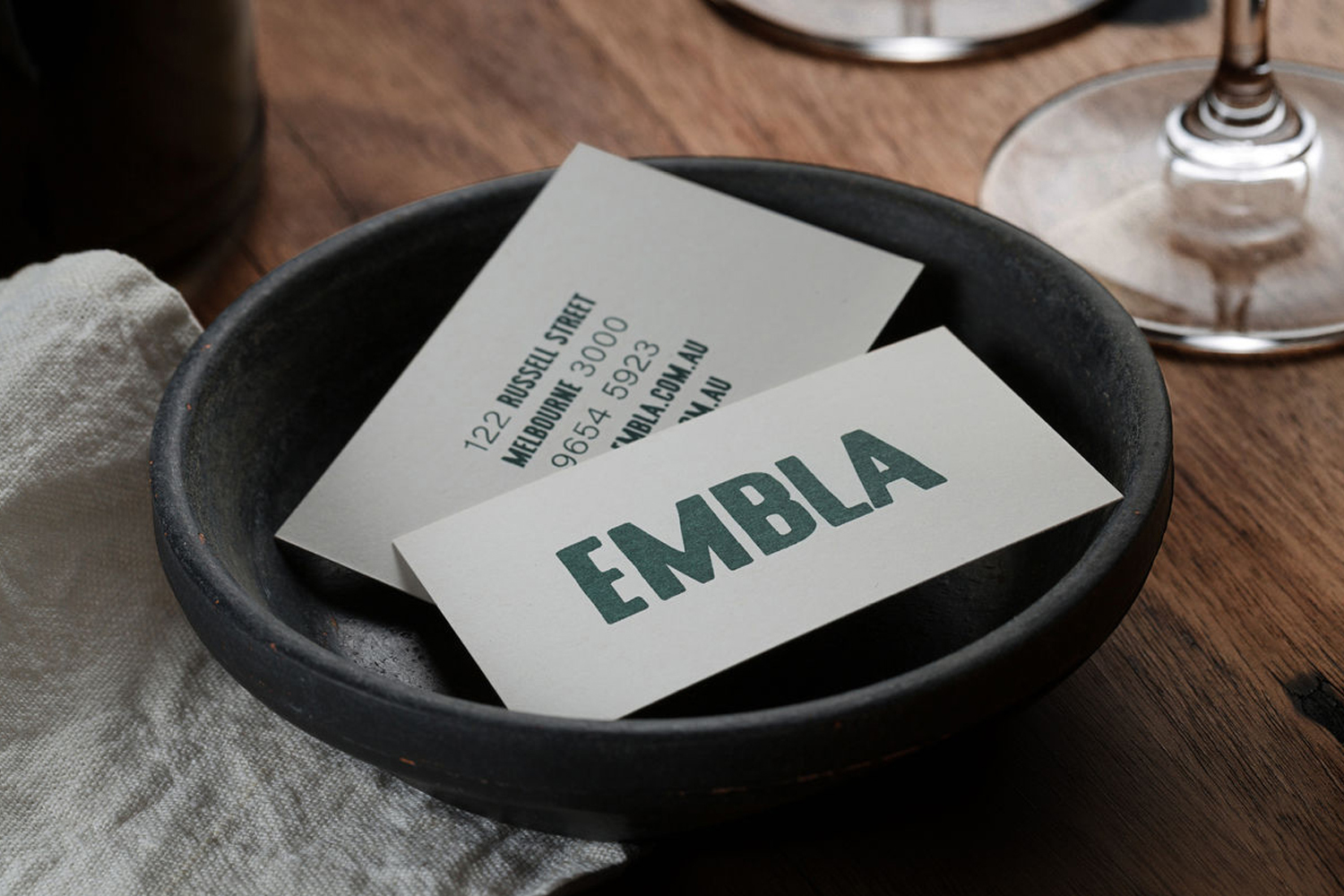 Embla restaurant business cards resting inside a dark ceramic bowl on a wooden table with wine glasses
