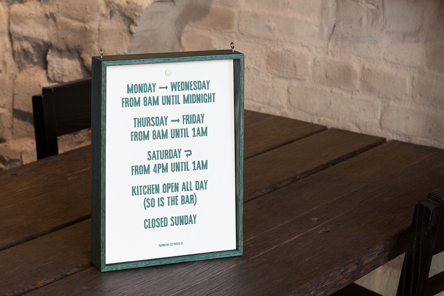 Framed sign showing operating hours resting on a dark wooden table against an exposed brick wall background.