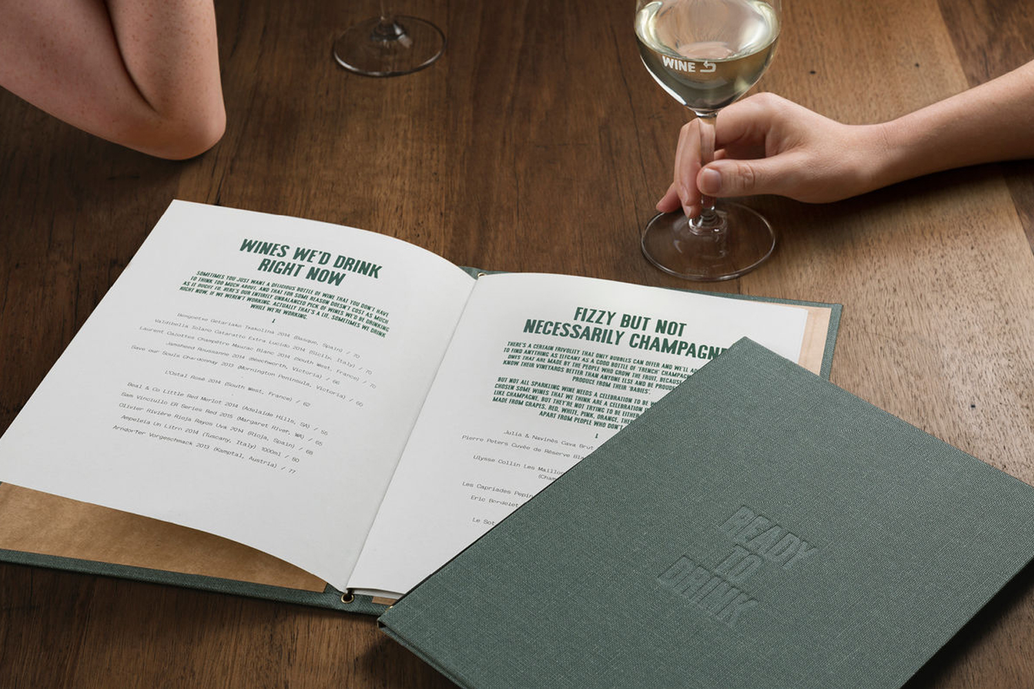 An open wine list menu on a wooden table with a hand holding a wine glass and a green bound cover bearing debossed text.