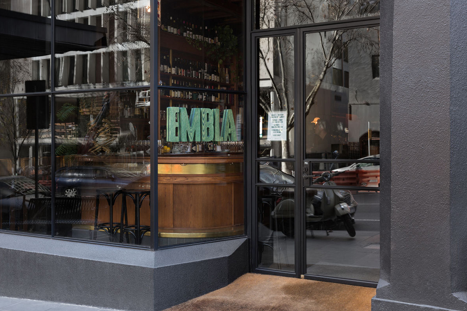 Exterior storefront view of Embla showing large glass windows, wooden bar interior, and turquoise EMBLA lettering signage.