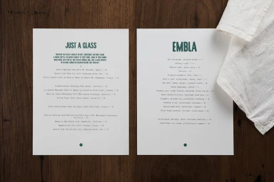 Two white paper menus with green typography resting on a dark wooden table beside a folded white linen napkin.