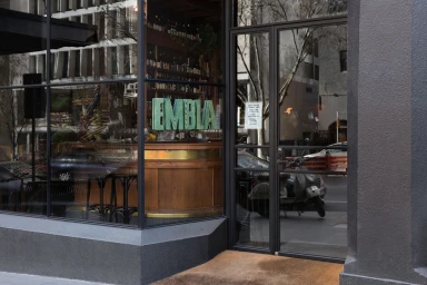 Exterior storefront view of Embla showing large glass windows, wooden bar interior, and turquoise EMBLA lettering signage.