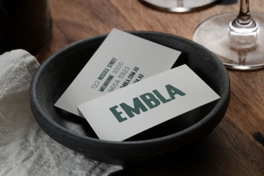 Embla restaurant business cards resting inside a dark ceramic bowl on a wooden table with wine glasses