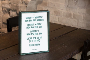 Framed sign showing operating hours resting on a dark wooden table against an exposed brick wall background.