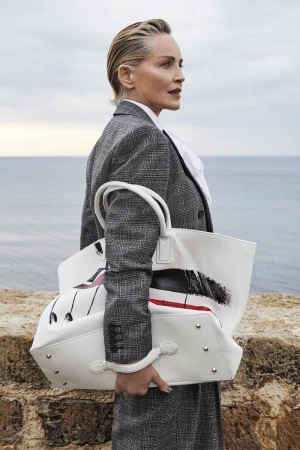 Sharon Stone wearing a grey plaid blazer holding a large white leather tote bag near the ocean for Antonio Marras