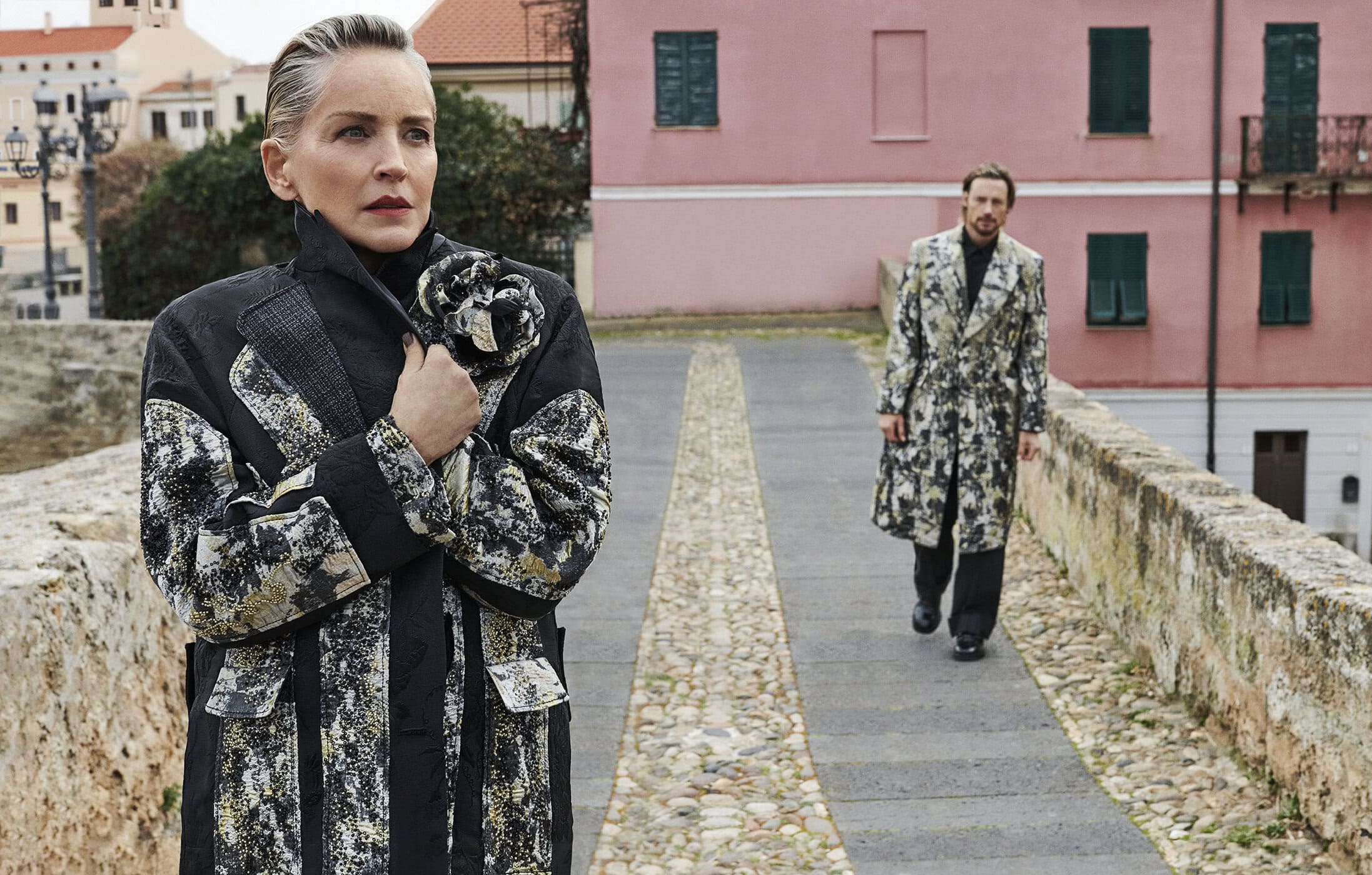 Sharon Stone in patterned coat in foreground with Gabriel Aubry walking behind her on stone walkway with pink building
