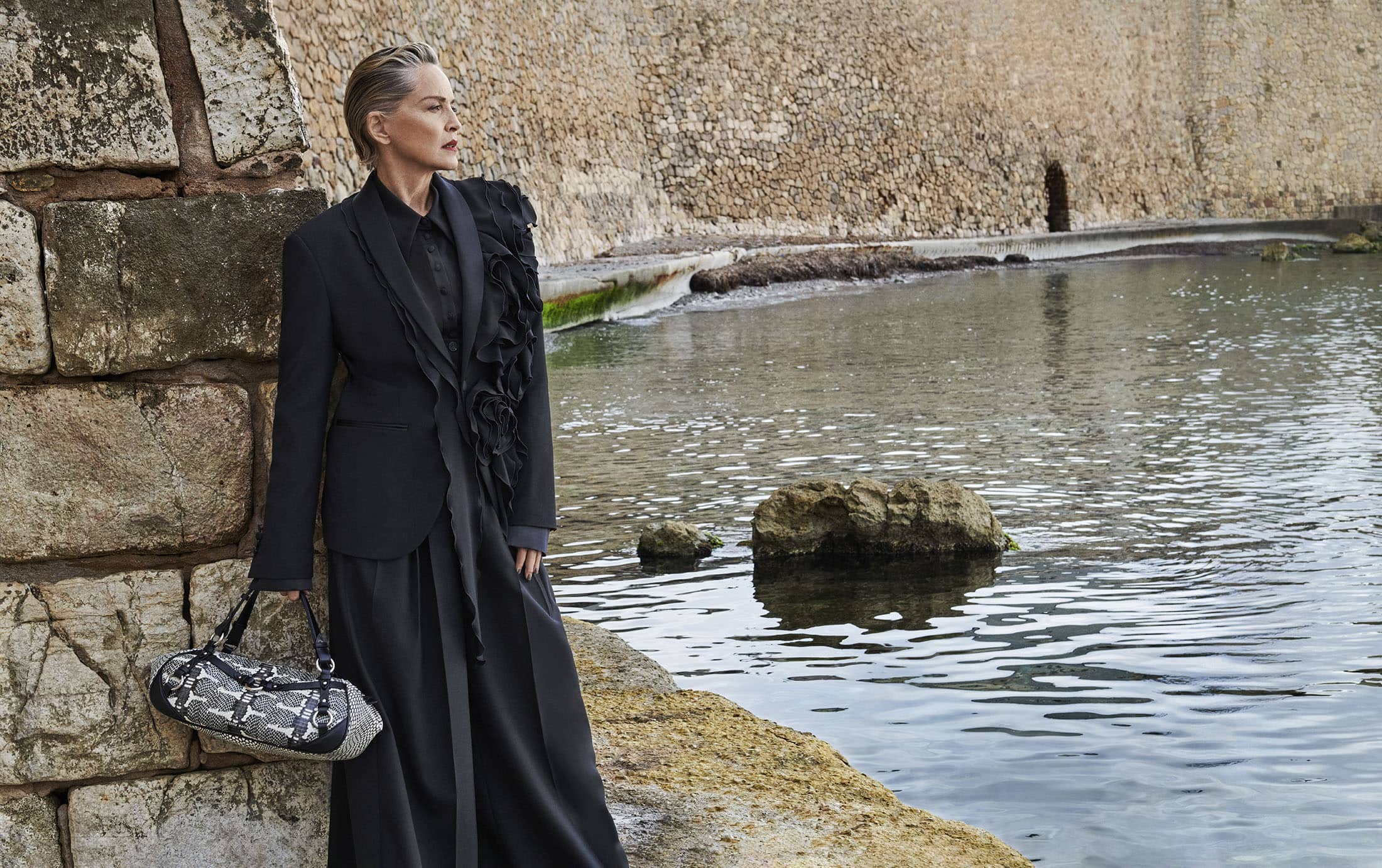 Sharon Stone wearing a black blazer and trousers holding a patterned handbag by a stone wall overlooking water