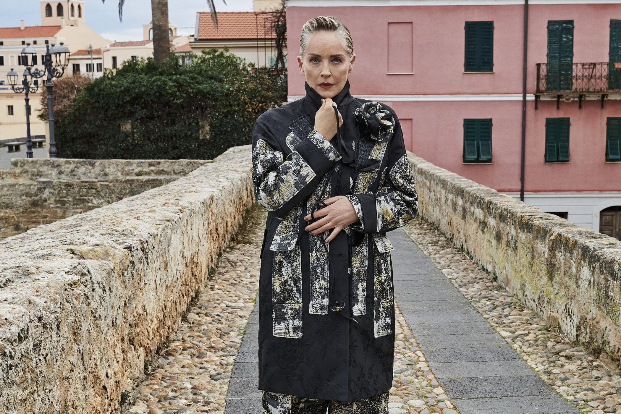 Sharon Stone standing on a stone bridge wearing a patterned black coat for the Antonio Marras Fall 2025 campaign.