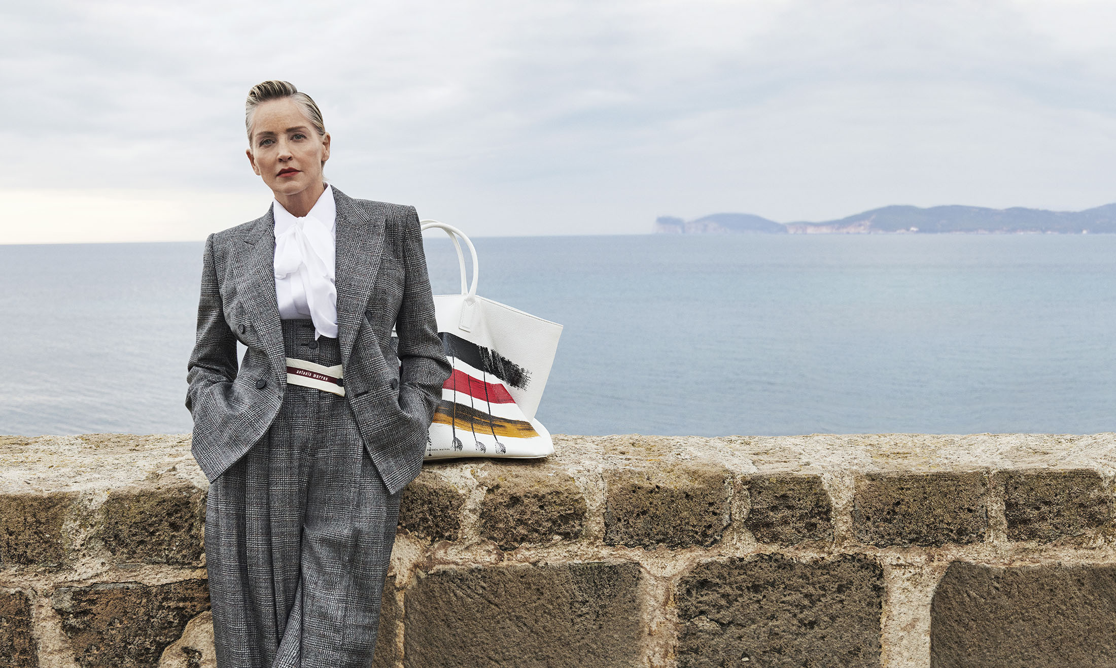 Sharon Stone in a grey plaid pantsuit leaning against a stone wall by the sea for Antonio Marras