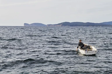 Man sitting in a small white wooden rowboat on the open sea with distant coastal hills under an overcast sky.