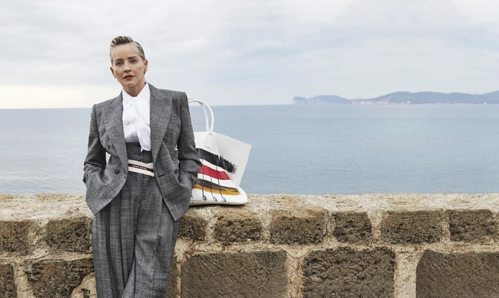 Sharon Stone in a grey plaid pantsuit leaning against a stone wall by the sea for Antonio Marras