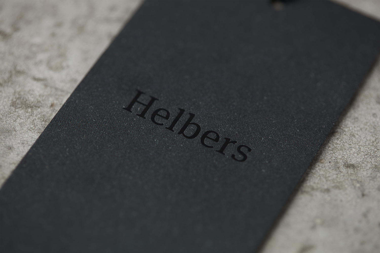 Black paper clothing tag with embossed Helbers brand logo resting on a textured concrete surface.